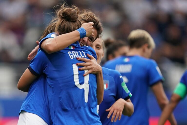 alt="Squadra di calcio femminile italiana unita sul campo da gioco, con le giocatrici che si abbracciano, di fronte a uno stadio con tifosi, a rappresentare la crescita e lo spirito collettivo del calcio femminile."