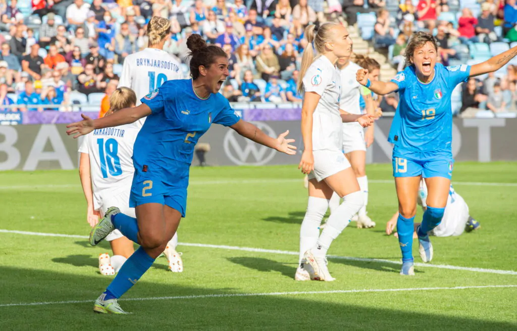 alt="Squadra di calcio femminile italiana in campo, vista di spalle, con le giocatrici abbracciate e unite, di fronte a uno stadio con tifosi, a rappresentare la crescita e lo spirito di squadra del calcio femminile in Italia."